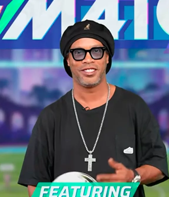 Ronaldinho Gaúcho segurando bola de futebol e logo atrás um estádio do jogo Rematch e a logo do jogo.
