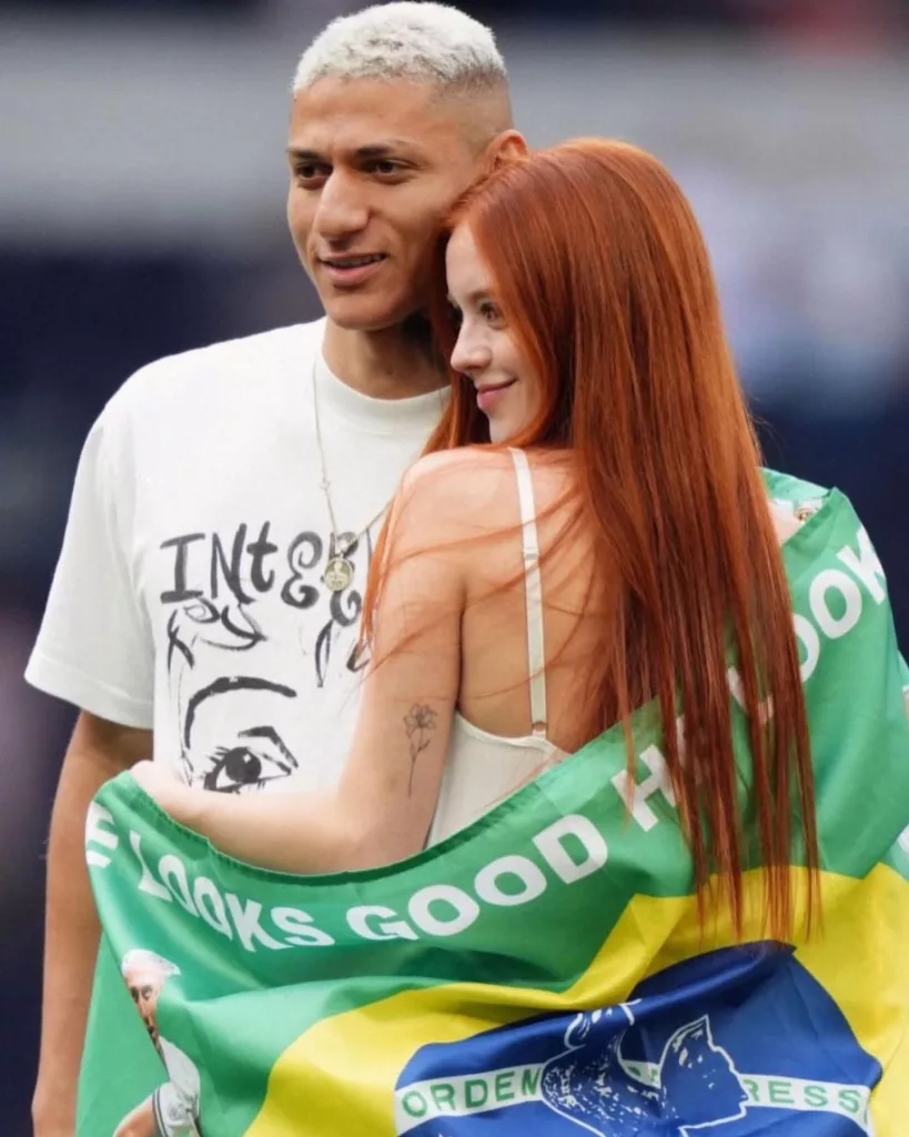Richarlison e Amanda abraçados e Amanda com bandeira do Brasil nas costas.
