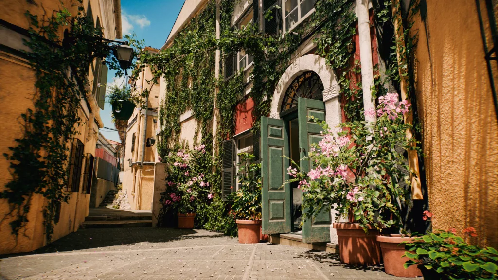 Viela com flores e vegetação geral com casas alaranjadas e céu azul.