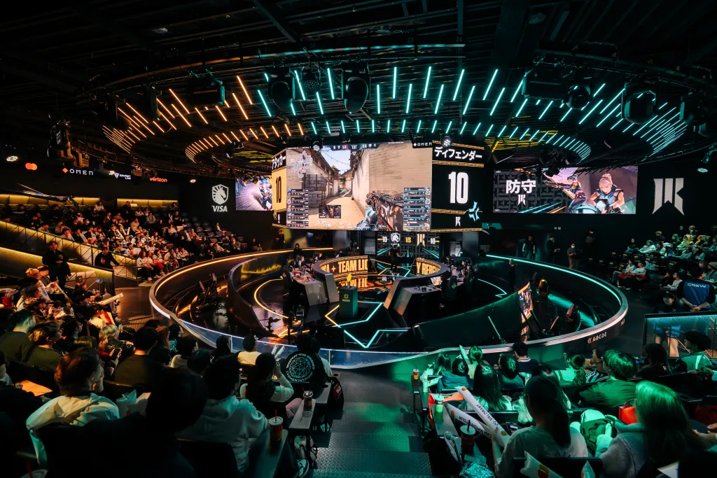 Foto de plateia em volta e jogadoras no centro em meio ao campeonato, além de telões no topo mostrando o jogo.