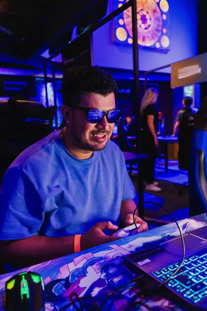 Homem de óculos escuros em frente a notebook com controle na mão e sorrindo.