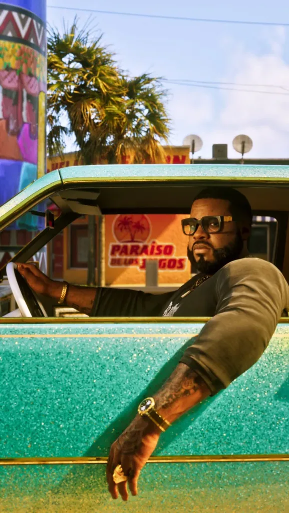 Personagem Boobie Ike em carro verde com muito brilho em frente a um local chamado Paraíso de los jugos. Está de óculos , correntes douradas e camiseta com mangas semi-cumpridas.